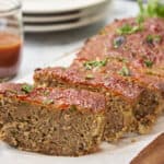 Juicy meatloaf topped with tomato sauce and garnished with chopped parsley on a wooden cutting board. A glass of ketchup and a stack of plates are visible in the background. Perfect comfort food for a family dinner.