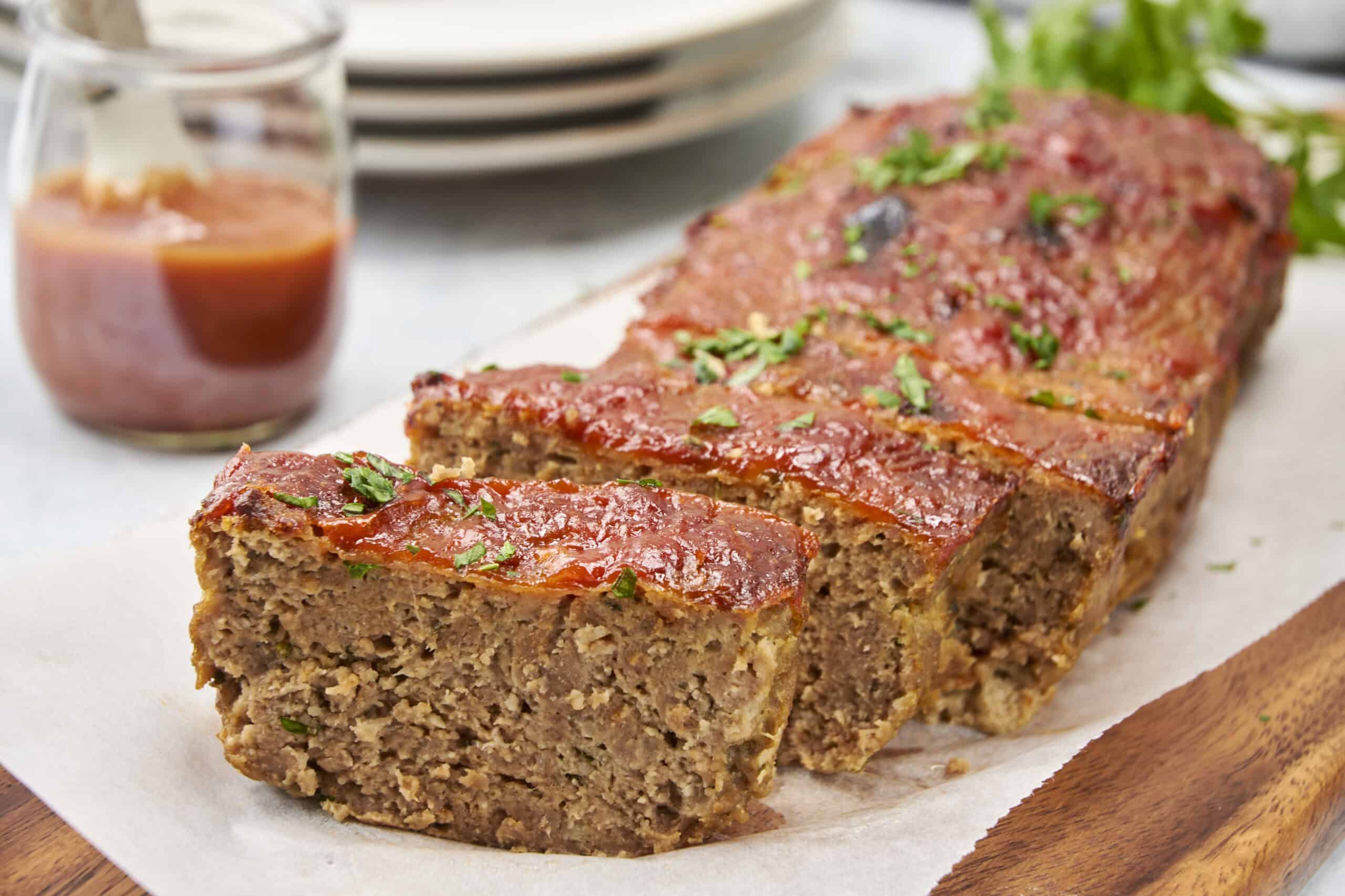 Juicy meatloaf topped with tomato sauce and garnished with chopped parsley on a wooden cutting board. A glass of ketchup and a stack of plates are visible in the background. Perfect comfort food for a family dinner.
