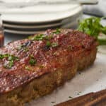 Juicy baked meatloaf garnished with chopped parsley on a wooden serving board, ready to serve with side dishes, with a small bowl of tomato ketchup and stacked white plates in the background.