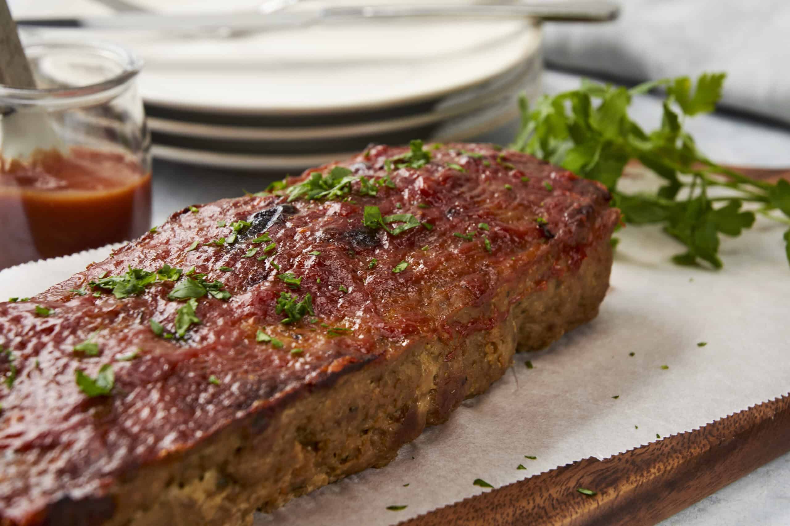 Juicy baked meatloaf garnished with chopped parsley on a wooden serving board, ready to serve with side dishes, with a small bowl of tomato ketchup and stacked white plates in the background.