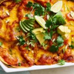 Golden baked enchiladas topped with melted cheese and fresh avocado, lime, and cilantro in a white ceramic baking dish, served with chopped green onions for a flavorful Mexican-inspired meal.
