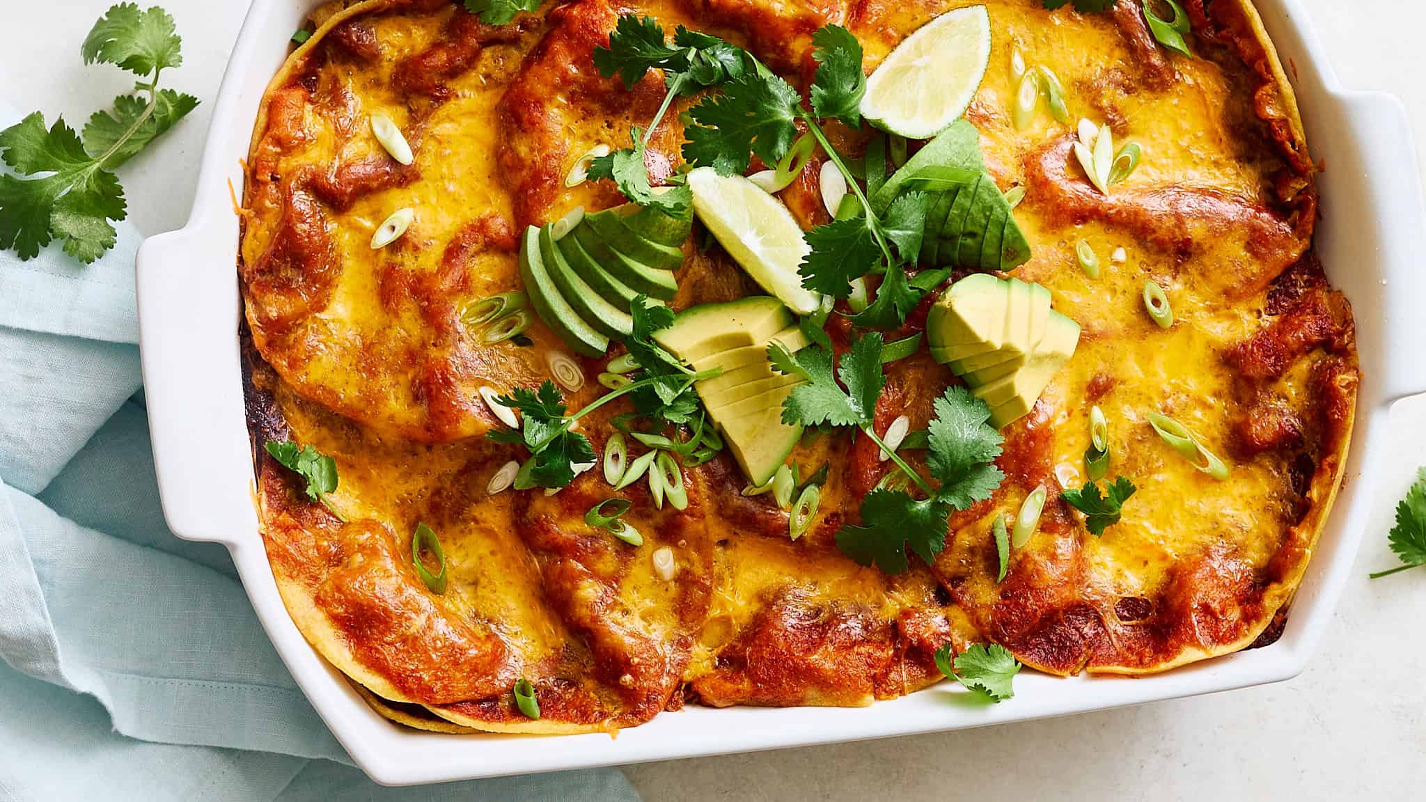 Golden baked enchiladas topped with melted cheese and fresh avocado, lime, and cilantro in a white ceramic baking dish, served with chopped green onions for a flavorful Mexican-inspired meal.