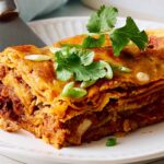 Crispy layered lasagna with ground beef, rich tomato sauce, and melted cheese, garnished with fresh cilantro and sliced green onions on a white decorative plate.