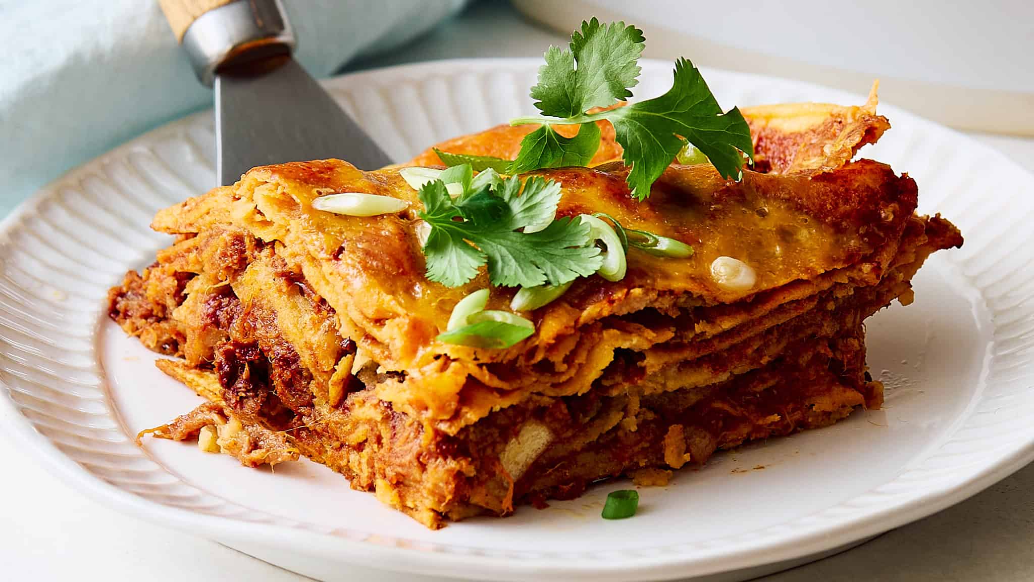 Crispy layered lasagna with ground beef, rich tomato sauce, and melted cheese, garnished with fresh cilantro and sliced green onions on a white decorative plate.