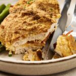 Golden baked chicken cordon bleu with crispy breadcrumb coating paired with fresh cooked asparagus on a ceramic plate. Perfect comfort food and easy homemade recipe for dinner or lunch.