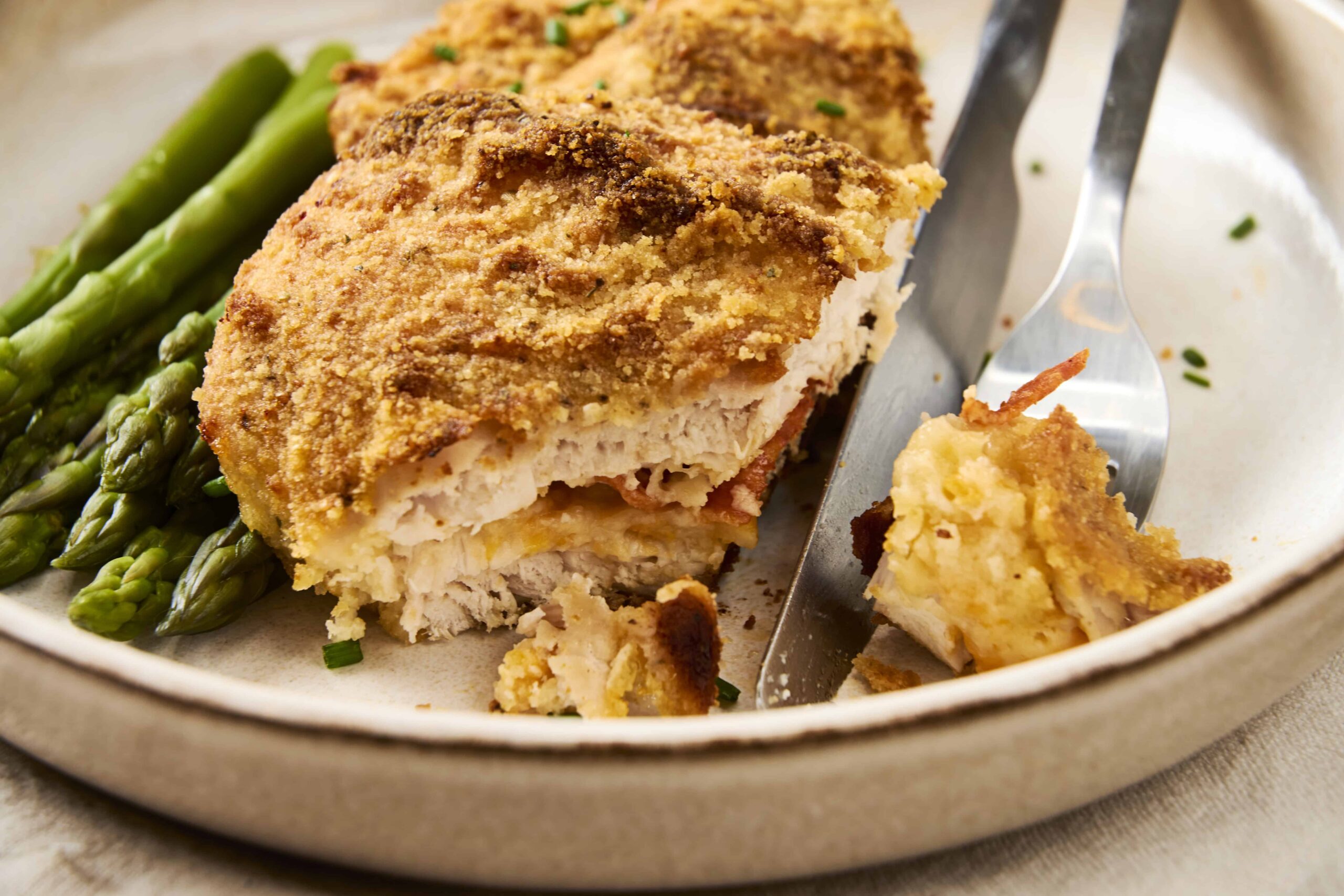 Golden baked chicken cordon bleu with crispy breadcrumb coating paired with fresh cooked asparagus on a ceramic plate. Perfect comfort food and easy homemade recipe for dinner or lunch.