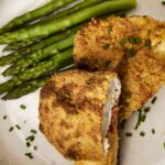 Crispy baked chicken cordon bleu with golden breadcrumb coating served with tender steamed asparagus on a white plate. Perfect comfort food with a crunchy crust and savory filling.