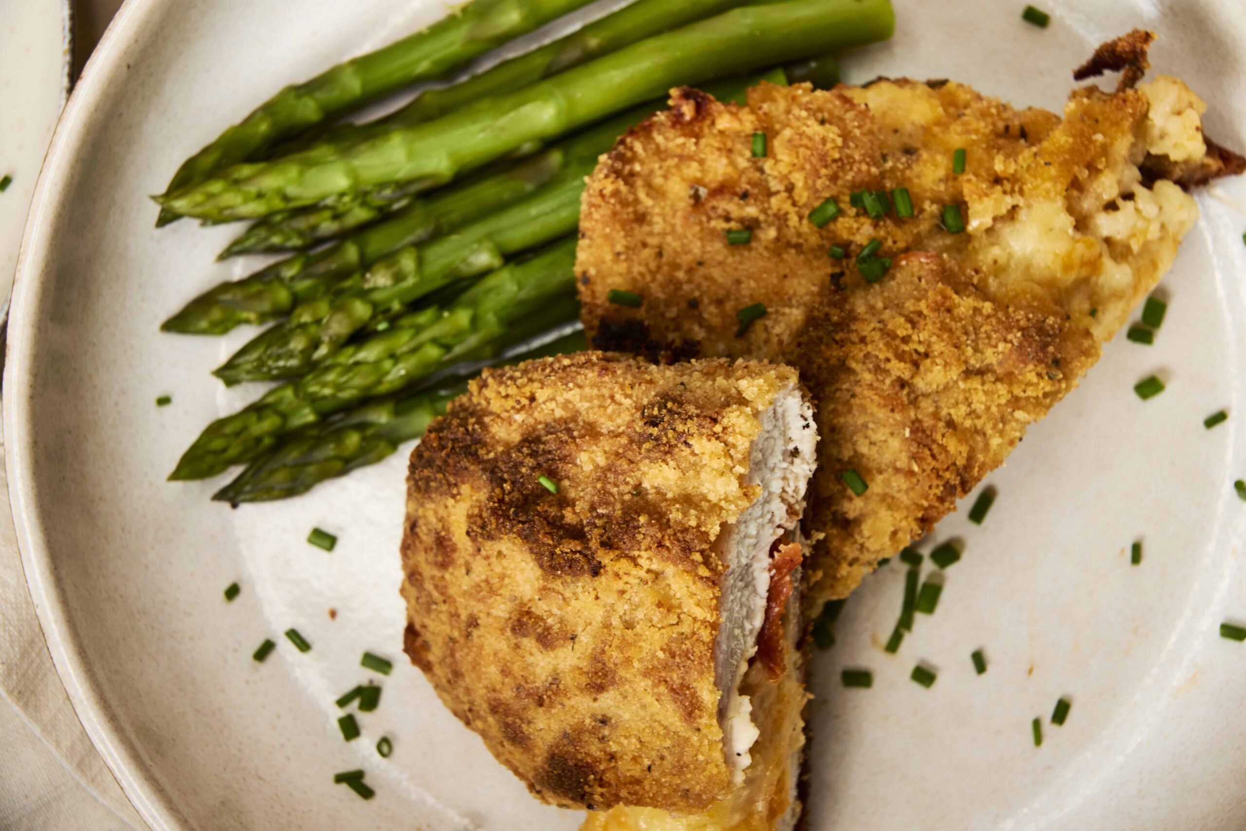 Crispy baked chicken cordon bleu with golden breadcrumb coating served with tender steamed asparagus on a white plate. Perfect comfort food with a crunchy crust and savory filling.