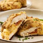 Crispy baked chicken cordon bleu with melted cheese and golden breadcrumb coating on a beige plate, garnished with chopped green onions, close-up, delicious chicken dish for dinner or lunch.