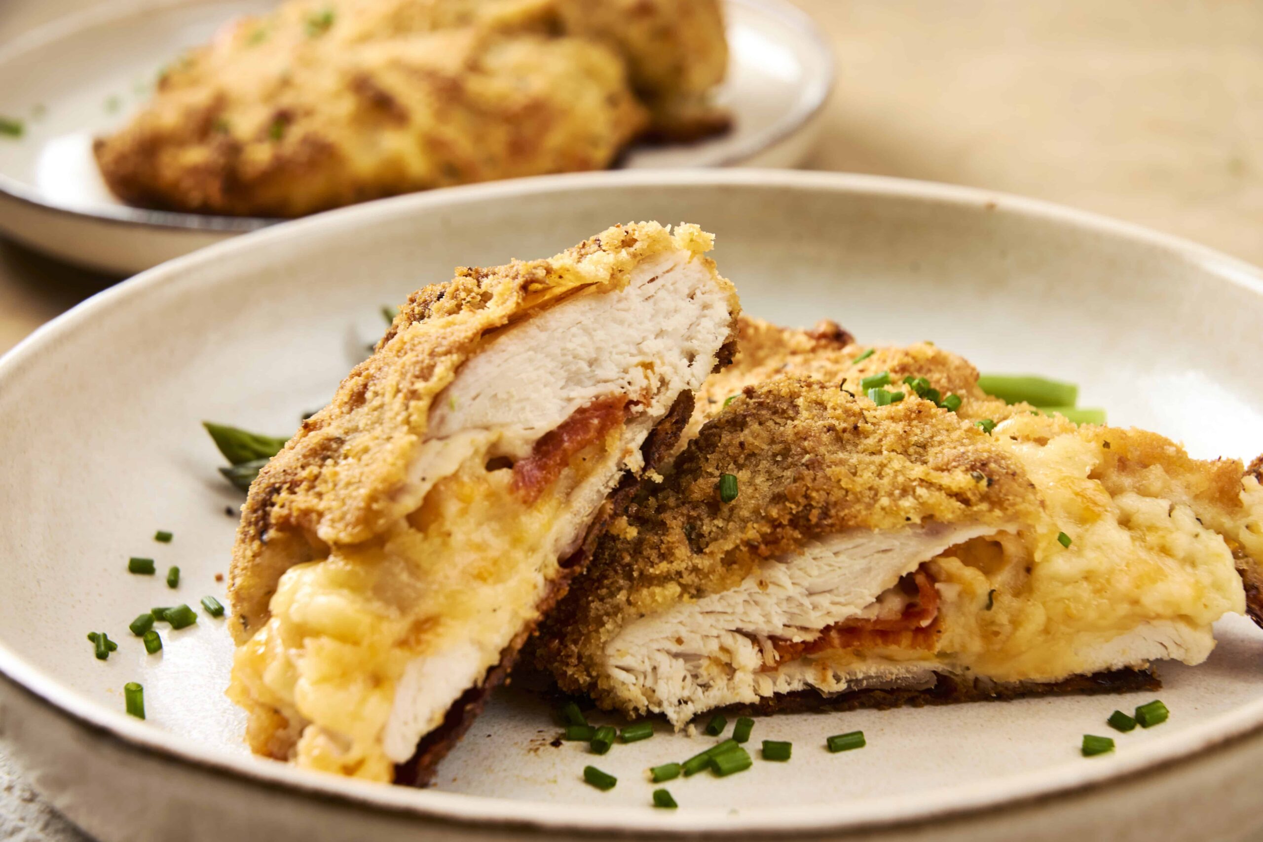 Crispy baked chicken cordon bleu with melted cheese and golden breadcrumb coating on a beige plate, garnished with chopped green onions, close-up, delicious chicken dish for dinner or lunch.