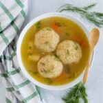 Fluffy chicken meatball soup with dill and vegetables in a white bowl on a gray background, garnished with fresh herbs, served with a wooden spoon and a checkered cloth, ideal for comforting homemade recipes.