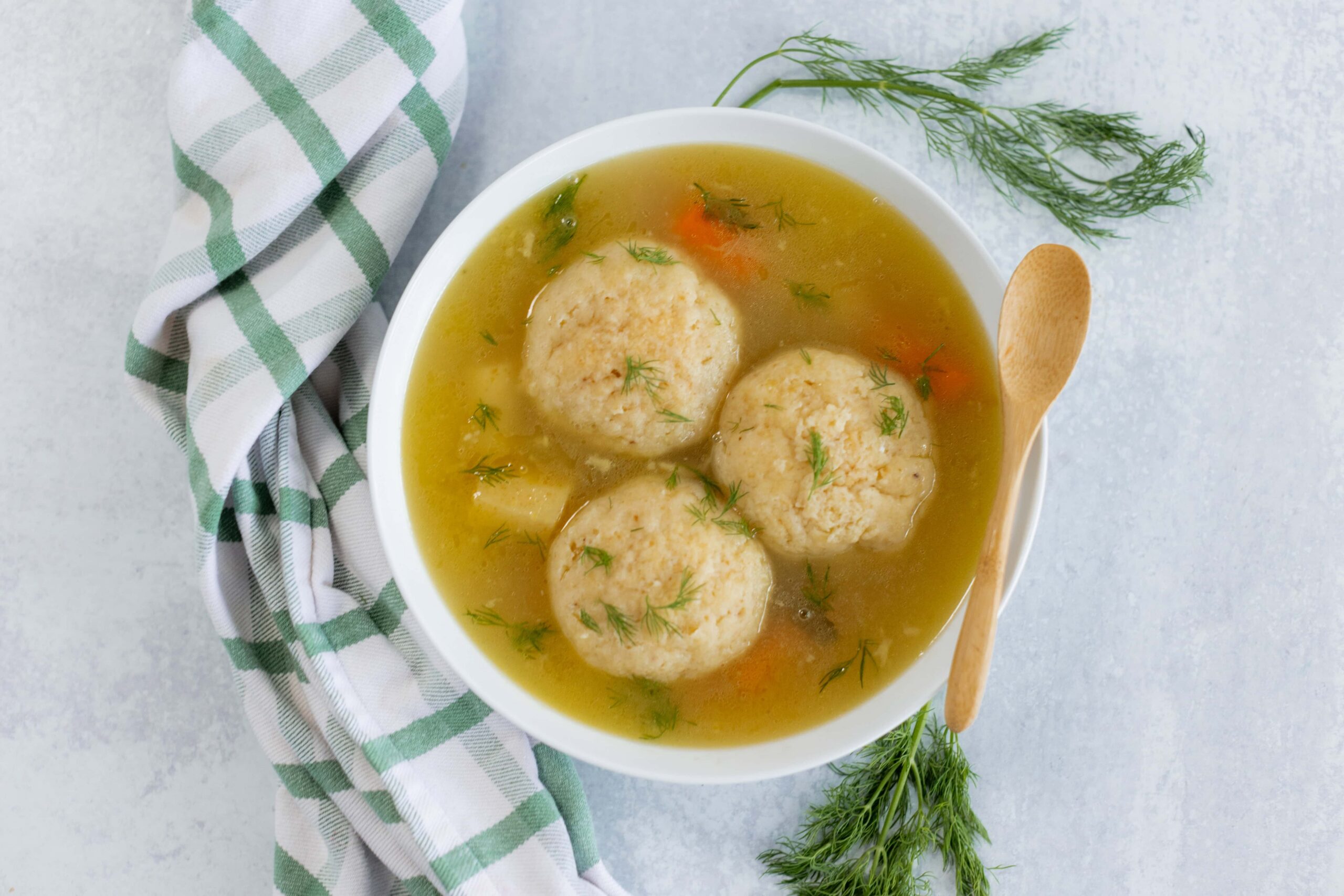 Fluffy chicken meatball soup with dill and vegetables in a white bowl on a gray background, garnished with fresh herbs, served with a wooden spoon and a checkered cloth, ideal for comforting homemade recipes.