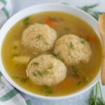 Fluffy chicken and dumplings soup with fresh herbs in a white bowl, served with a wooden spoon on a light background, perfect for cozy comfort food.