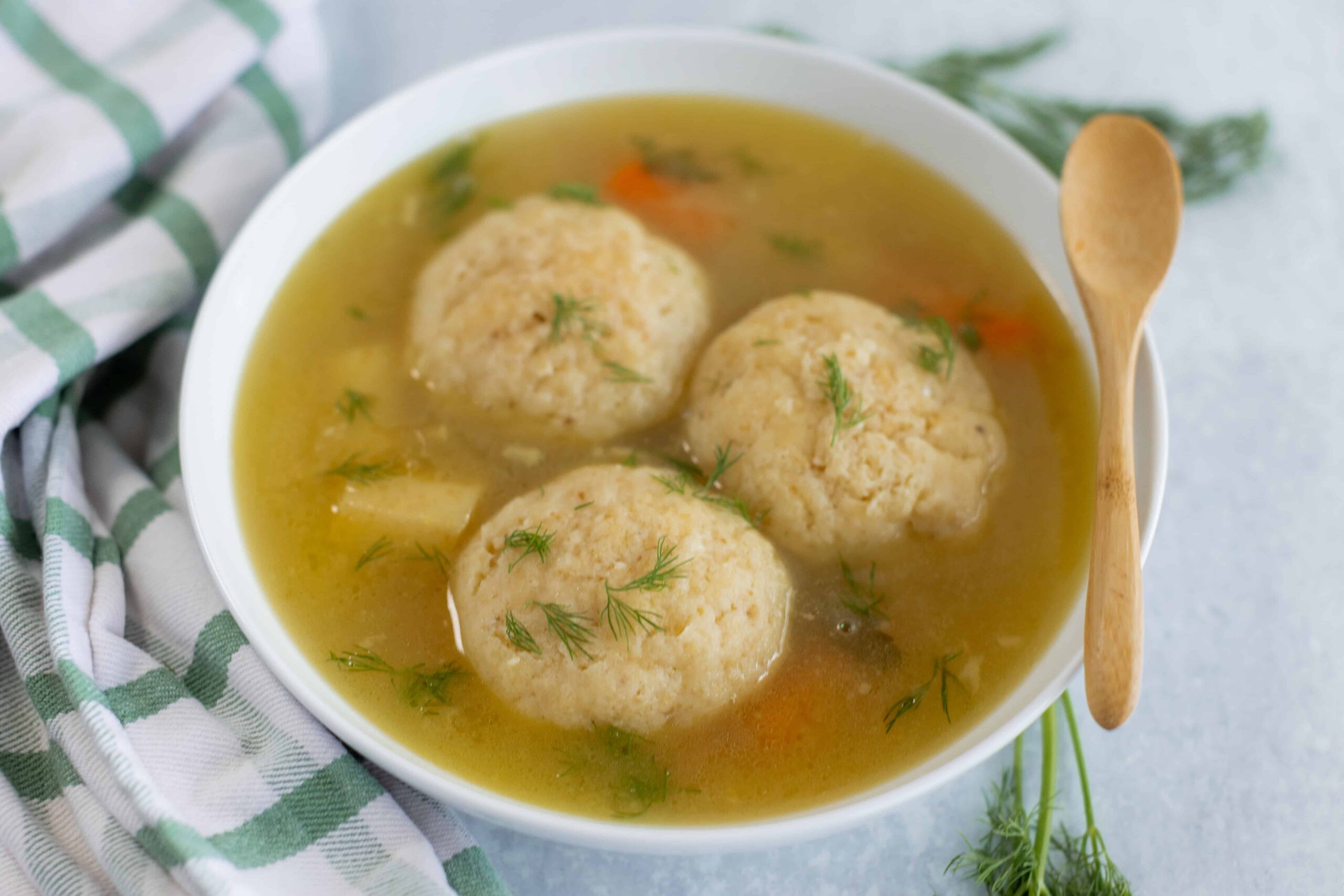 Fluffy chicken and dumplings soup with fresh herbs in a white bowl, served with a wooden spoon on a light background, perfect for cozy comfort food.