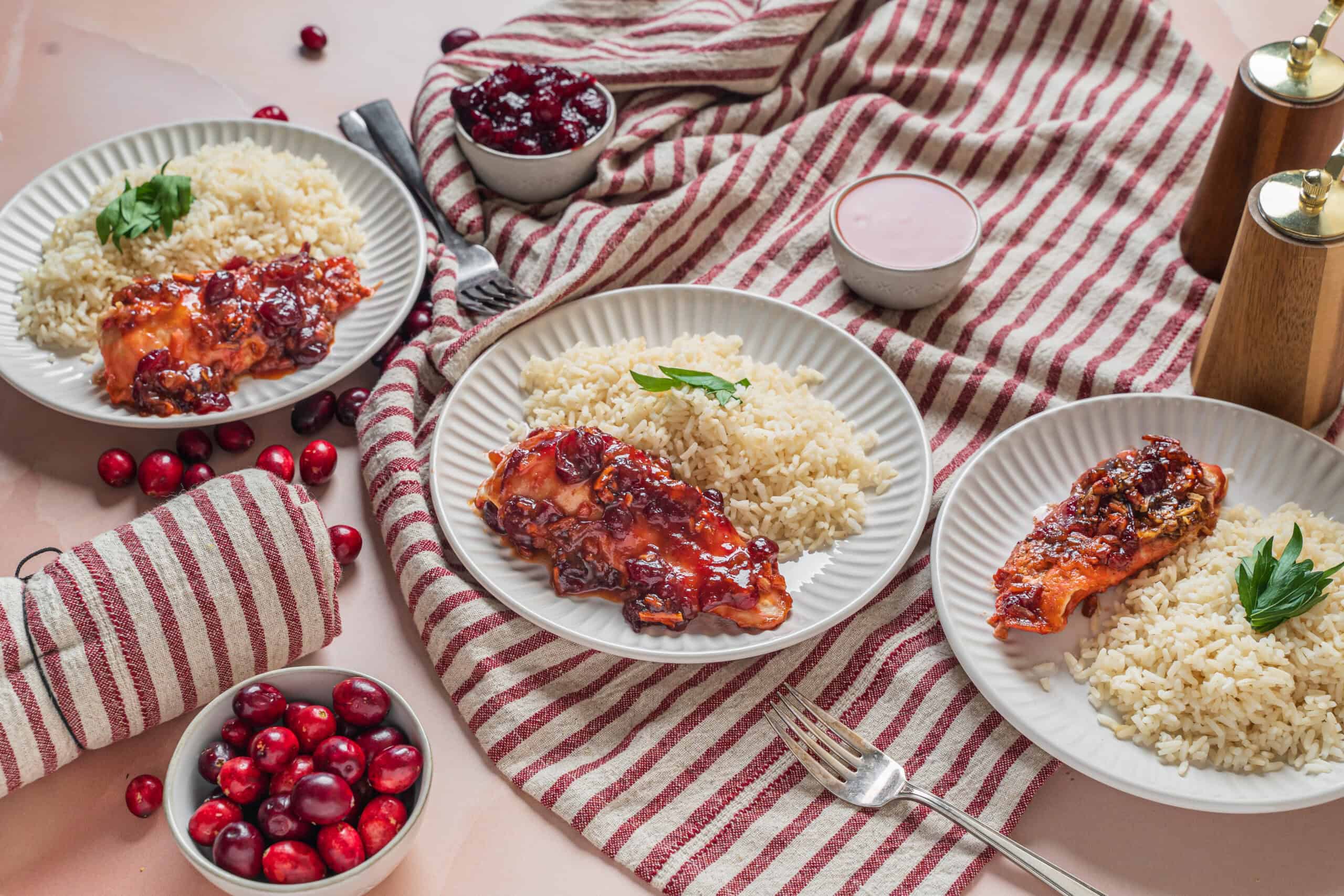 Juicy baked chicken thighs with cranberry sauce served with rice, garnished with fresh herbs on white plates, surrounded by cranberry bowls, cutlery, and rustic seasoning bottles for a cozy, festive meal.