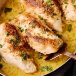 Juicy baked chicken breast with herbs served in a creamy sauce, garnished with fresh parsley, in a rustic skillet over dark wooden background.