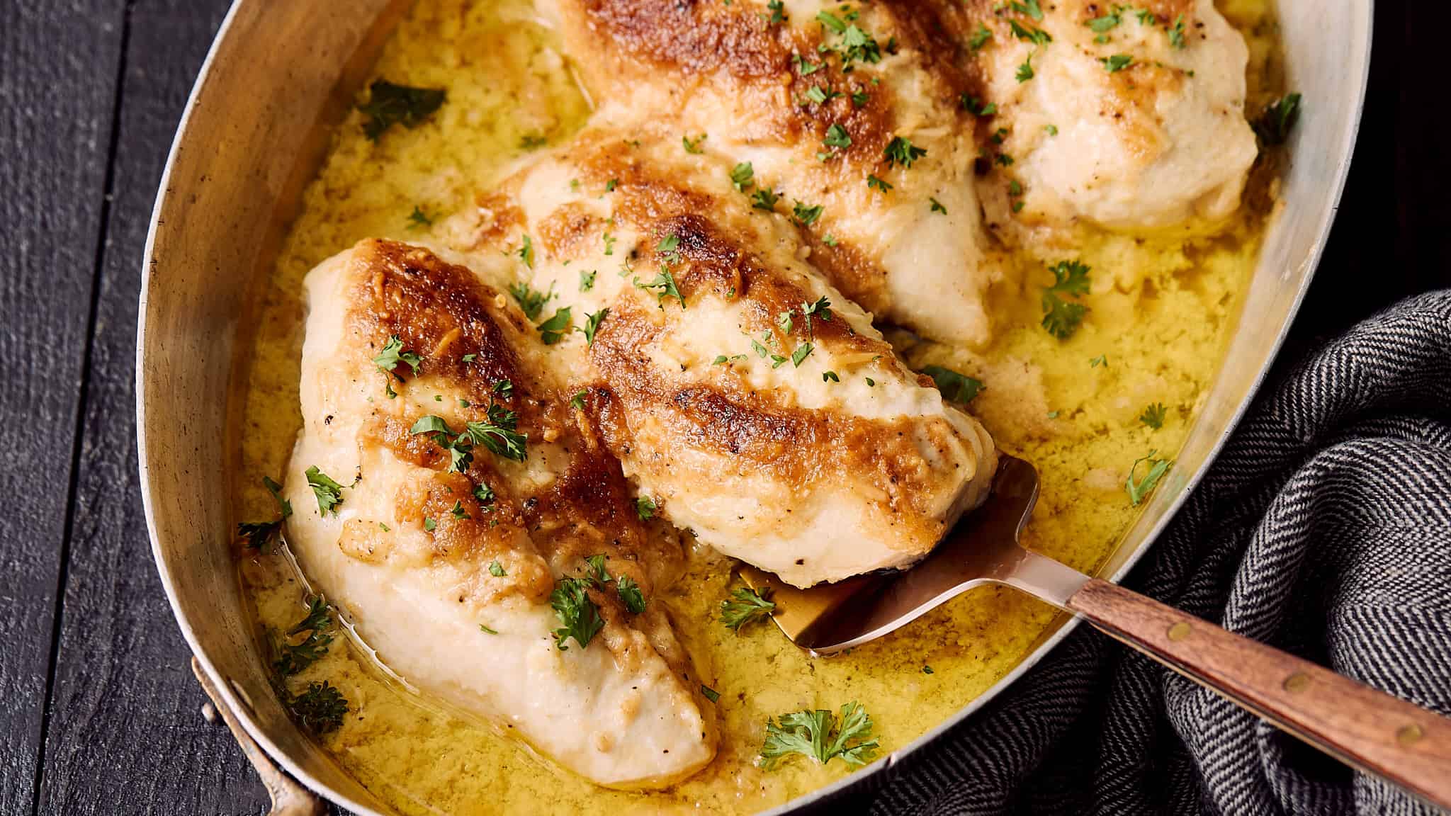 Juicy baked chicken breast with herbs served in a creamy sauce, garnished with fresh parsley, in a rustic skillet over dark wooden background.
