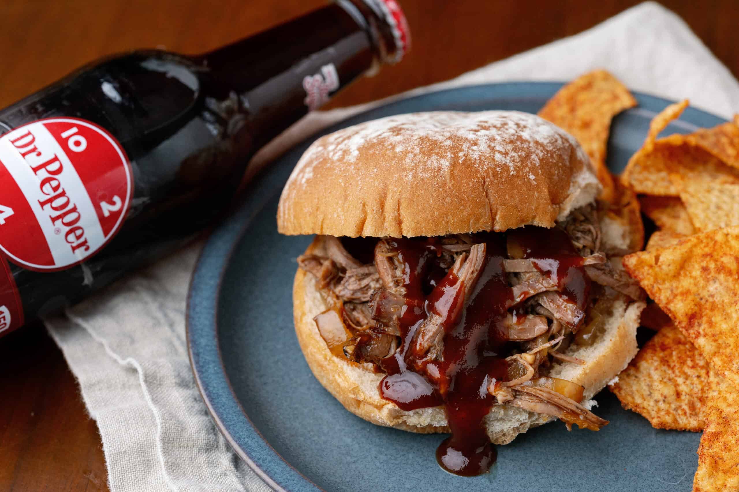 Juicy pulled pork sandwich with barbecue sauce on a blue plate, served with crispy tortilla chips, accompanied by an ice-cold bottle of Dr. Pepper soda, perfect for American comfort food meals.