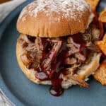 Tender shredded beef BBQ sandwich with smoky sauce, served on a toasted bun with crispy tortilla chips on a blue plate. Perfect comfort food meal with savory flavors and crunchy sides.
