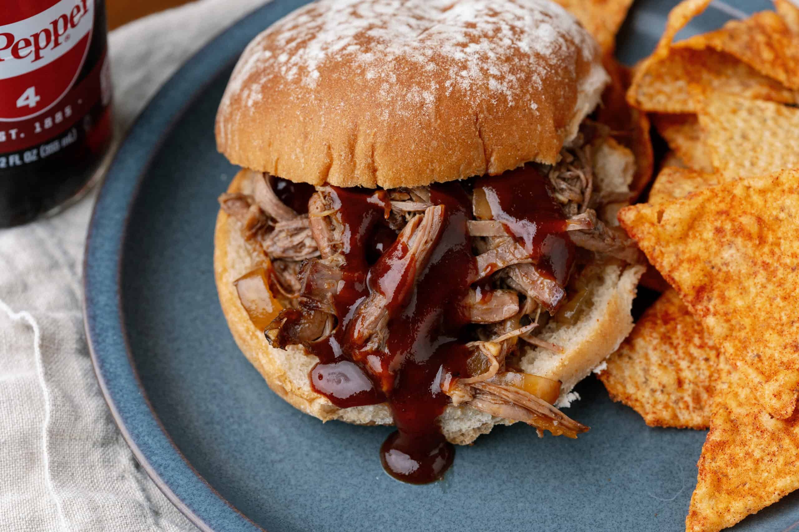 Tender shredded beef BBQ sandwich with smoky sauce, served on a toasted bun with crispy tortilla chips on a blue plate. Perfect comfort food meal with savory flavors and crunchy sides.