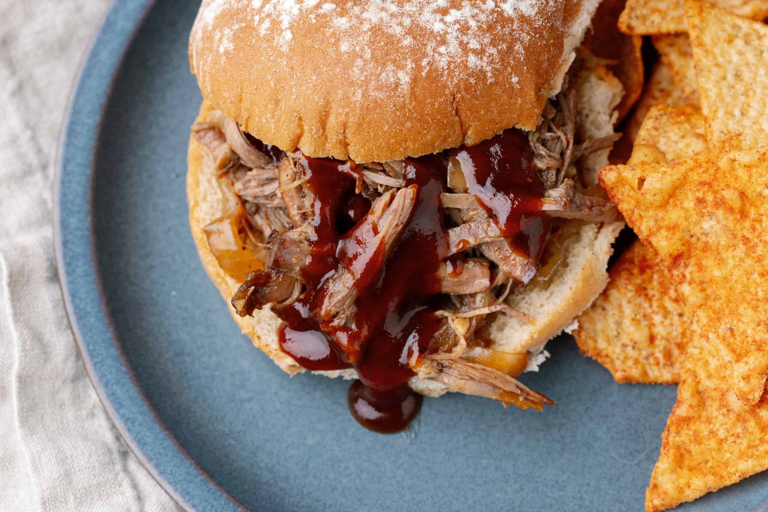 Succulent pulled pork sandwich with barbecue sauce served on a bun, paired with crispy tortilla chips on a blue plate, perfect for a delicious meal.