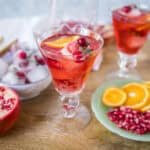 Refreshing pomegranate and orange summer cocktail with ice, garnished with fresh fruit slices and herbs, served in elegant glasses.