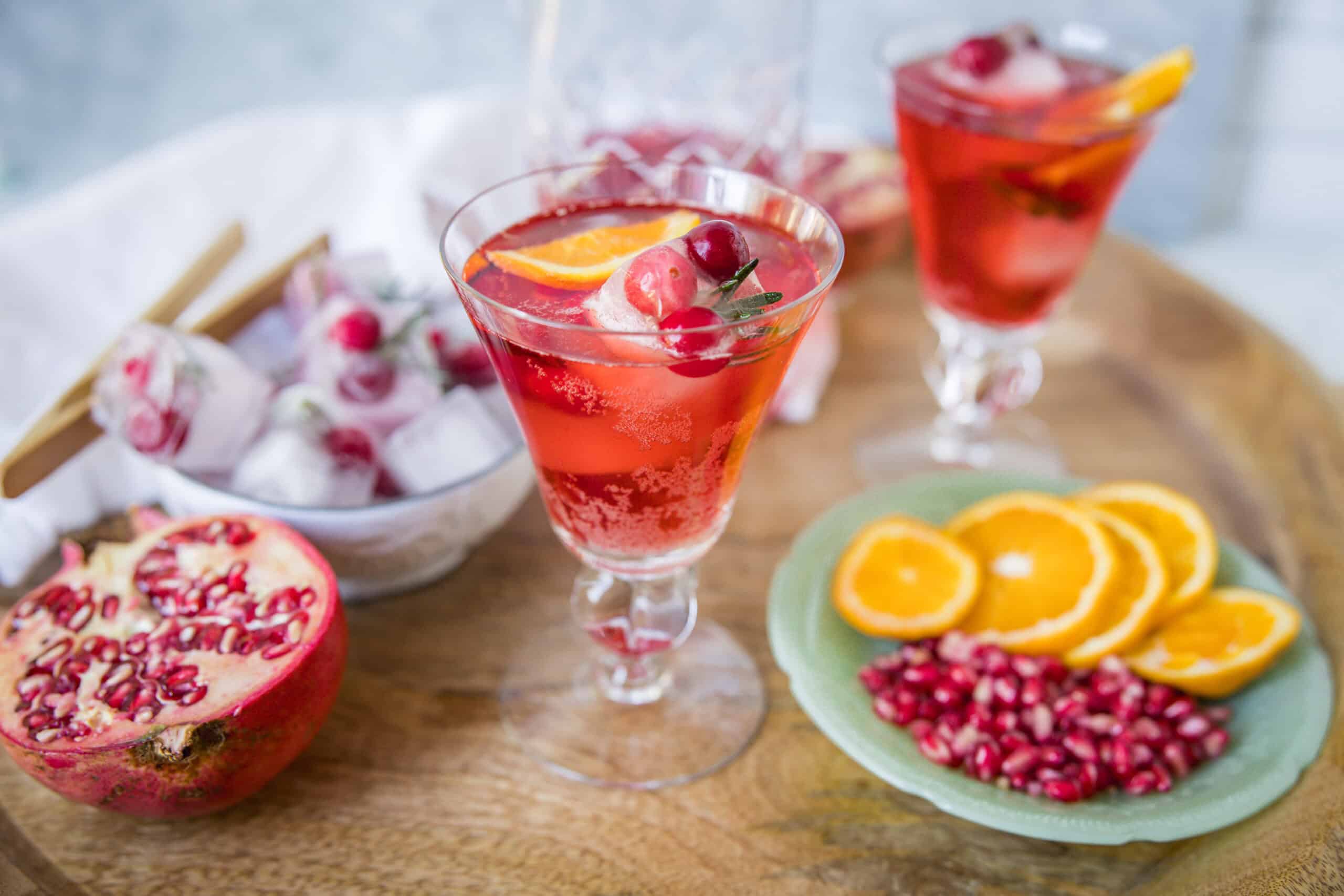 Refreshing pomegranate and orange summer cocktail with ice, garnished with fresh fruit slices and herbs, served in elegant glasses.