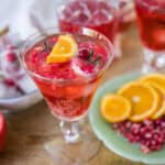 Refreshing pomegranate and orange cocktail garnished with fresh fruit slices, served in an elegant glass with a backdrop of pomegranate seeds and additional cocktail glasses.