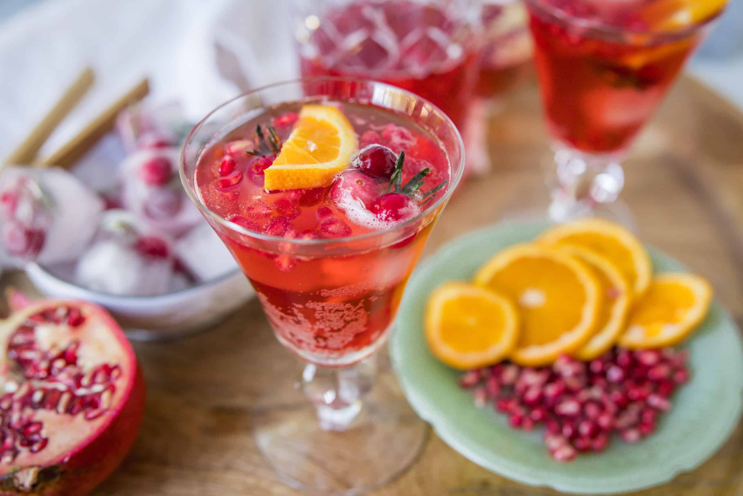 Refreshing pomegranate and orange cocktail garnished with fresh fruit slices, served in an elegant glass with a backdrop of pomegranate seeds and additional cocktail glasses.