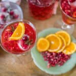Bright pomegranate and orange cocktail garnished with fresh fruit slices and pomegranate seeds, served in elegant glassware on a rustic wooden table. Perfect for summer parties and festive celebrations.