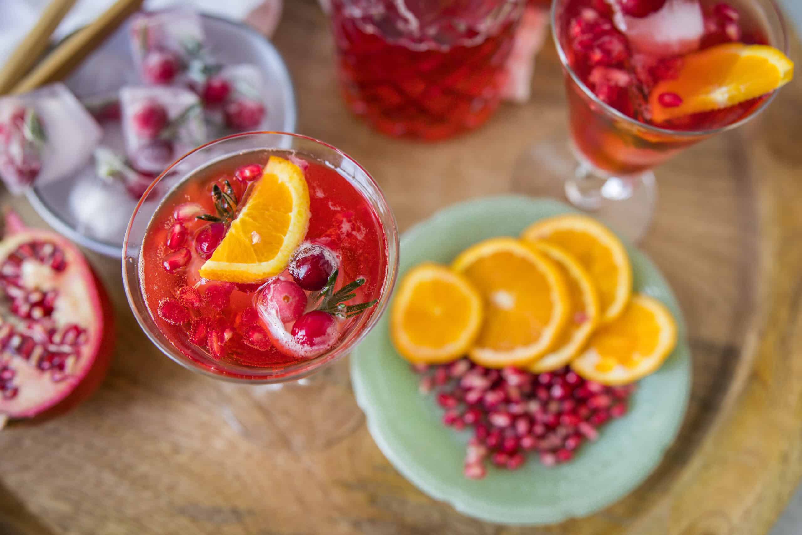 Bright pomegranate and orange cocktail garnished with fresh fruit slices and pomegranate seeds, served in elegant glassware on a rustic wooden table. Perfect for summer parties and festive celebrations.