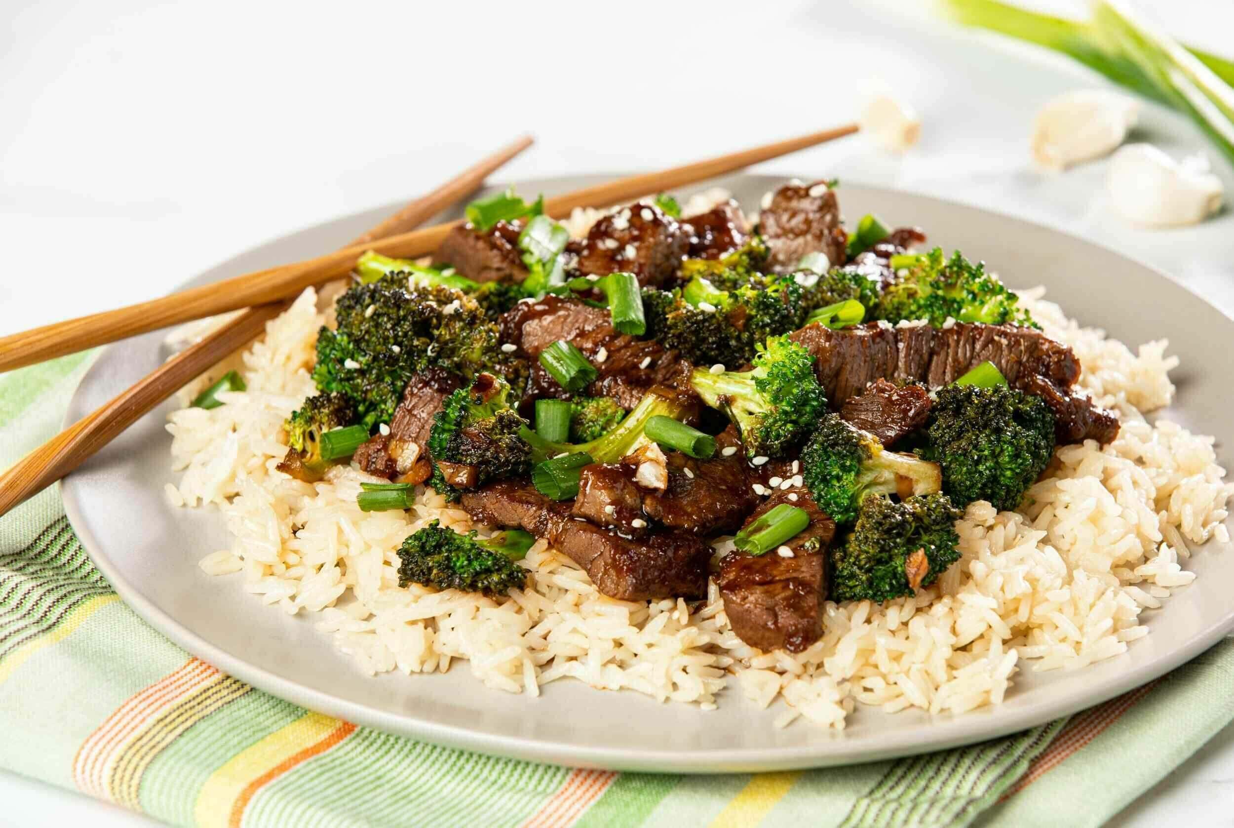 Savoury beef and broccoli stir-fry served over fluffy white rice on a white plate with chopsticks, garnished with sesame seeds and chopped green onions, perfect for a quick Asian-inspired meal.
