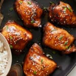Succulent glazed chicken thighs garnished with sesame seeds and green onions on a black serving platter.