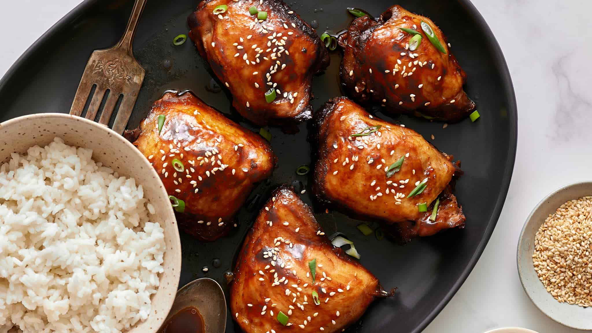 Succulent glazed chicken thighs garnished with sesame seeds and green onions on a black serving platter.