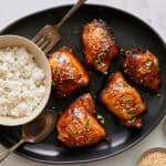 Succulent glazed chicken wings garnished with sesame seeds and chopped green onions served with a bowl of fluffy white rice and dipping sauces on a black platter.