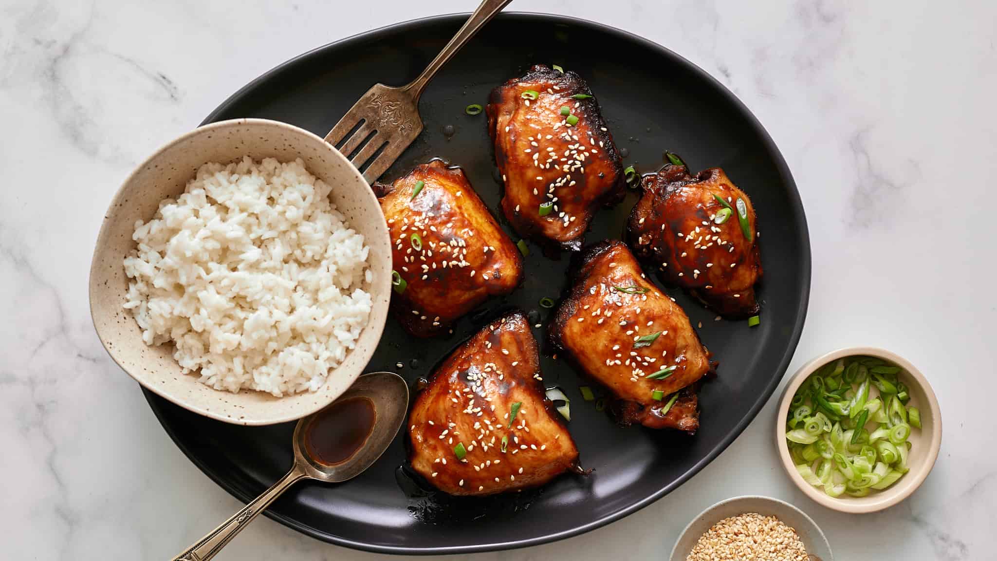 Succulent glazed chicken wings garnished with sesame seeds and chopped green onions served with a bowl of fluffy white rice and dipping sauces on a black platter.