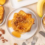 Golden brown French toast topped with chopped pecans served with syrup and fresh bananas on a light-colored table.