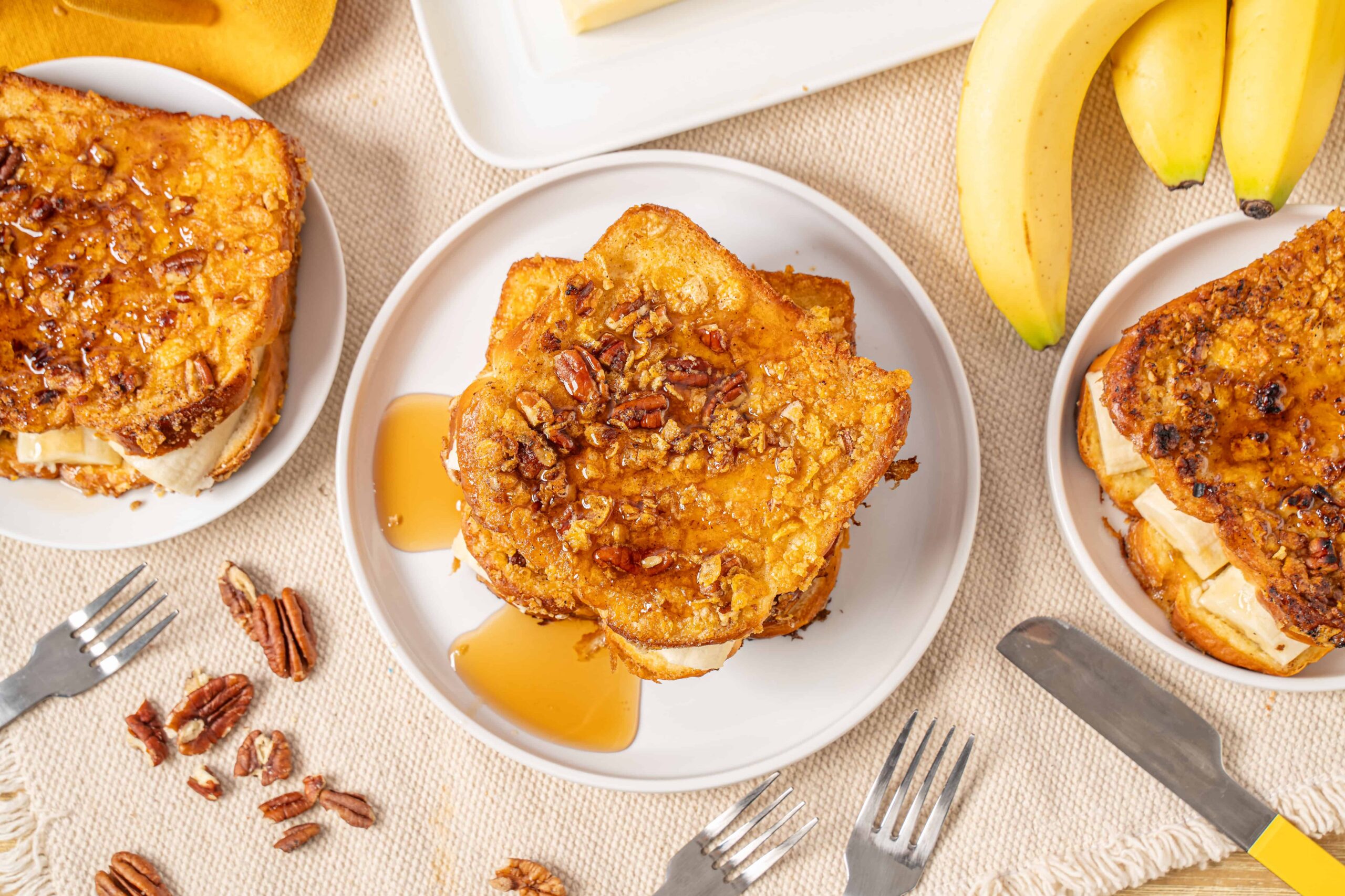 Golden brown French toast topped with chopped pecans served with syrup and fresh bananas on a light-colored table.
