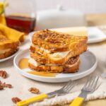 Golden caramelized banana French toast sandwiches with pecans, served with syrup and butter on a white plate. Perfect breakfast or brunch recipe for banana lovers.