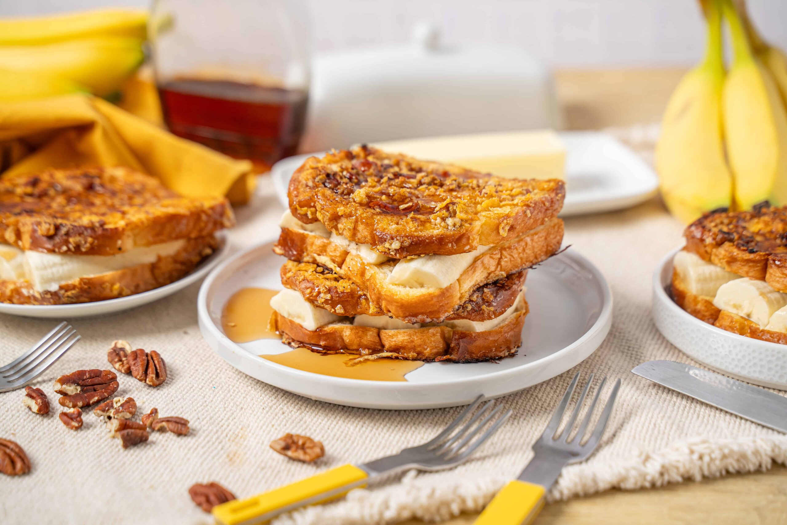 Golden caramelized banana French toast sandwiches with pecans, served with syrup and butter on a white plate. Perfect breakfast or brunch recipe for banana lovers.