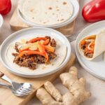 Savory beef taco with colorful bell peppers, onions, and soft flour tortillas on white plates, fresh ginger root on a beige surface, highlighting delicious homemade Mexican cuisine.