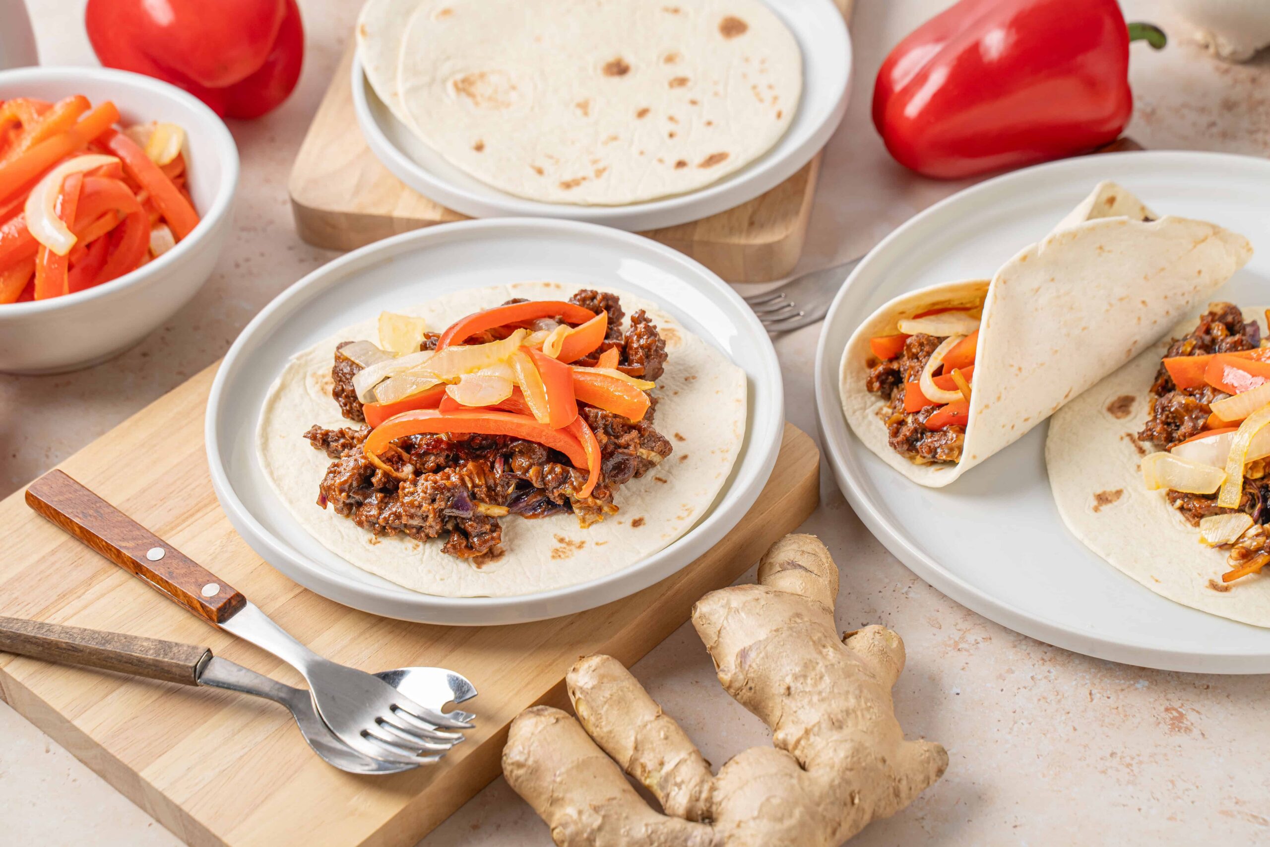 Savory beef taco with colorful bell peppers, onions, and soft flour tortillas on white plates, fresh ginger root on a beige surface, highlighting delicious homemade Mexican cuisine.