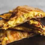 Golden scrambled eggs breakfast quesadilla filled with cheese and bacon, sliced in half, served on a dark plate. Perfect for a savory breakfast or brunch with crispy exterior and melty cheese inside.