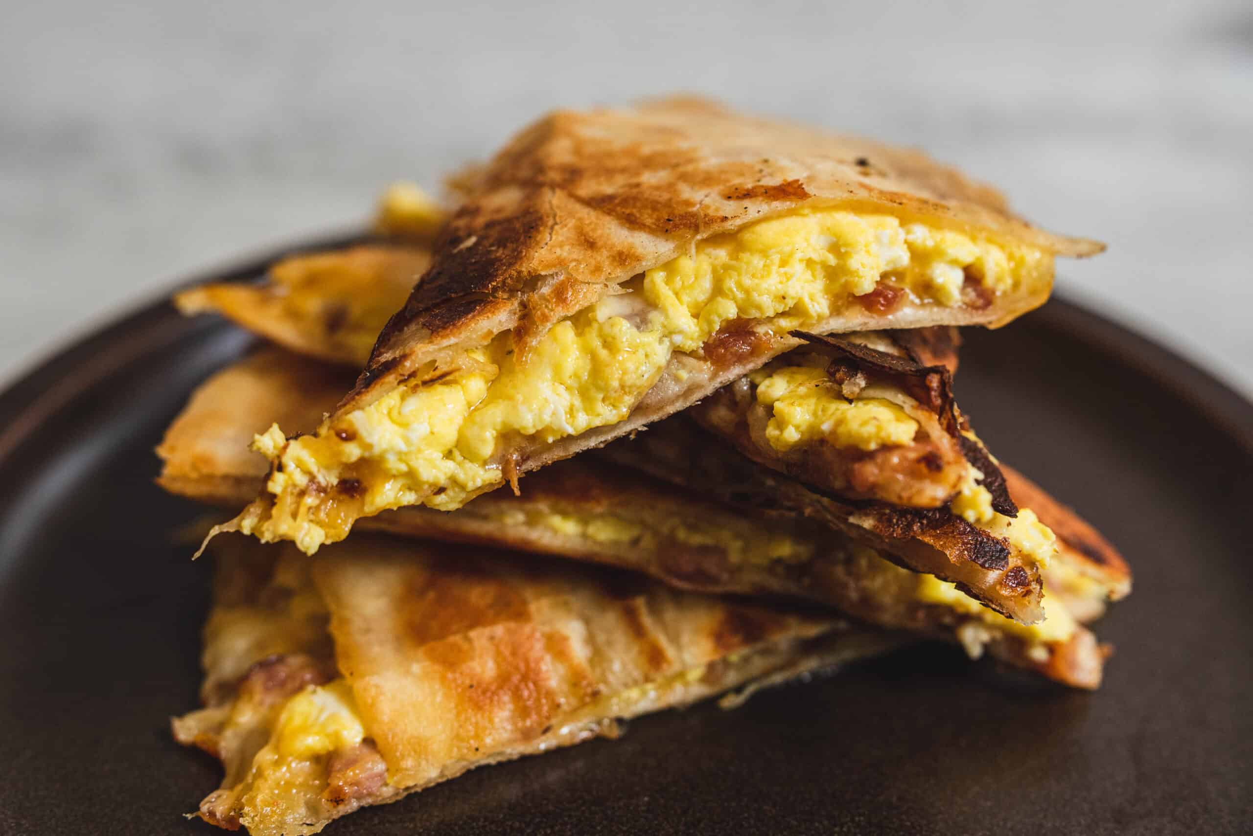 Golden scrambled eggs breakfast quesadilla filled with cheese and bacon, sliced in half, served on a dark plate. Perfect for a savory breakfast or brunch with crispy exterior and melty cheese inside.