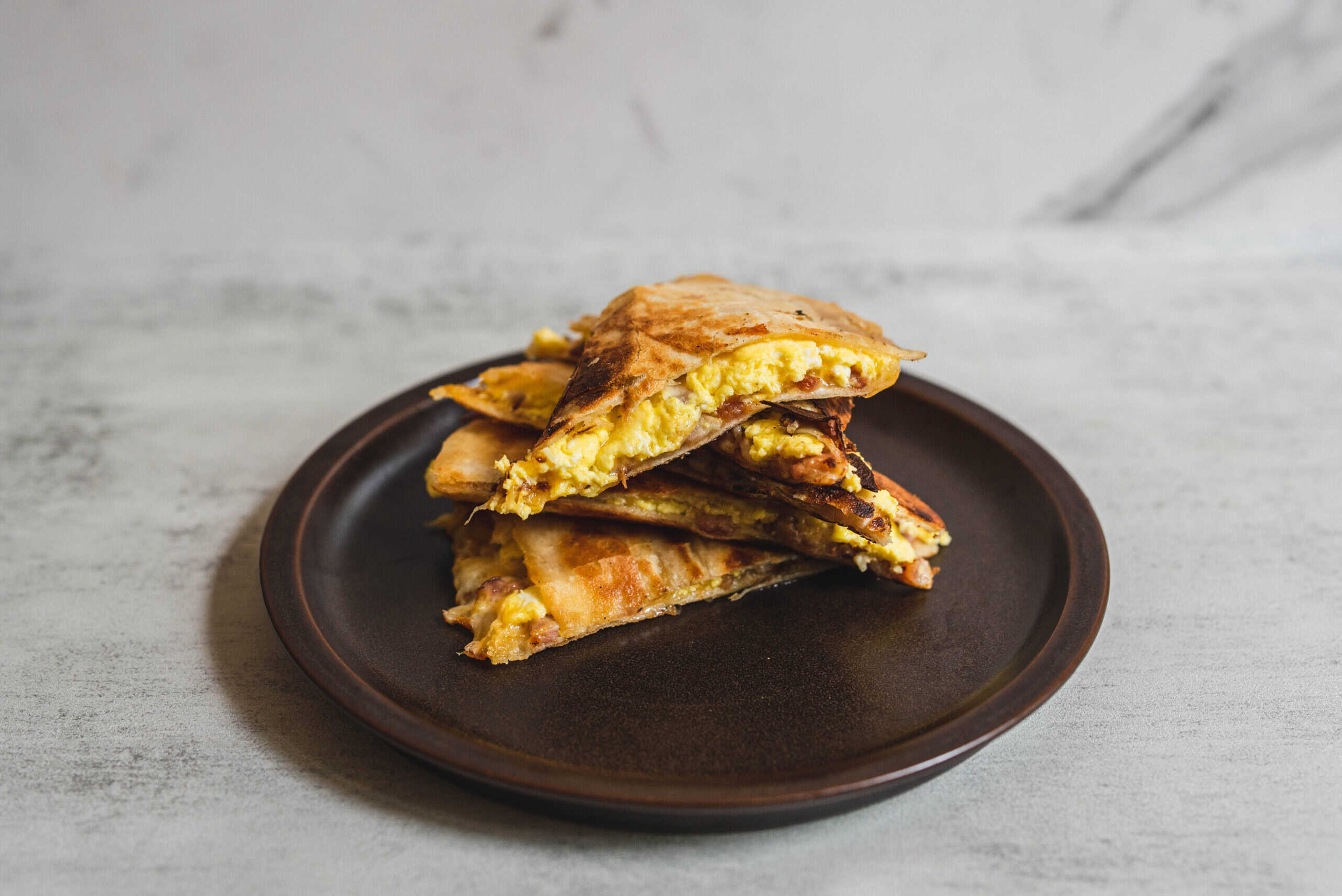 Crispy breakfast quesadilla with scrambled eggs, cheese, and bacon on a dark plate against a rustic gray background.