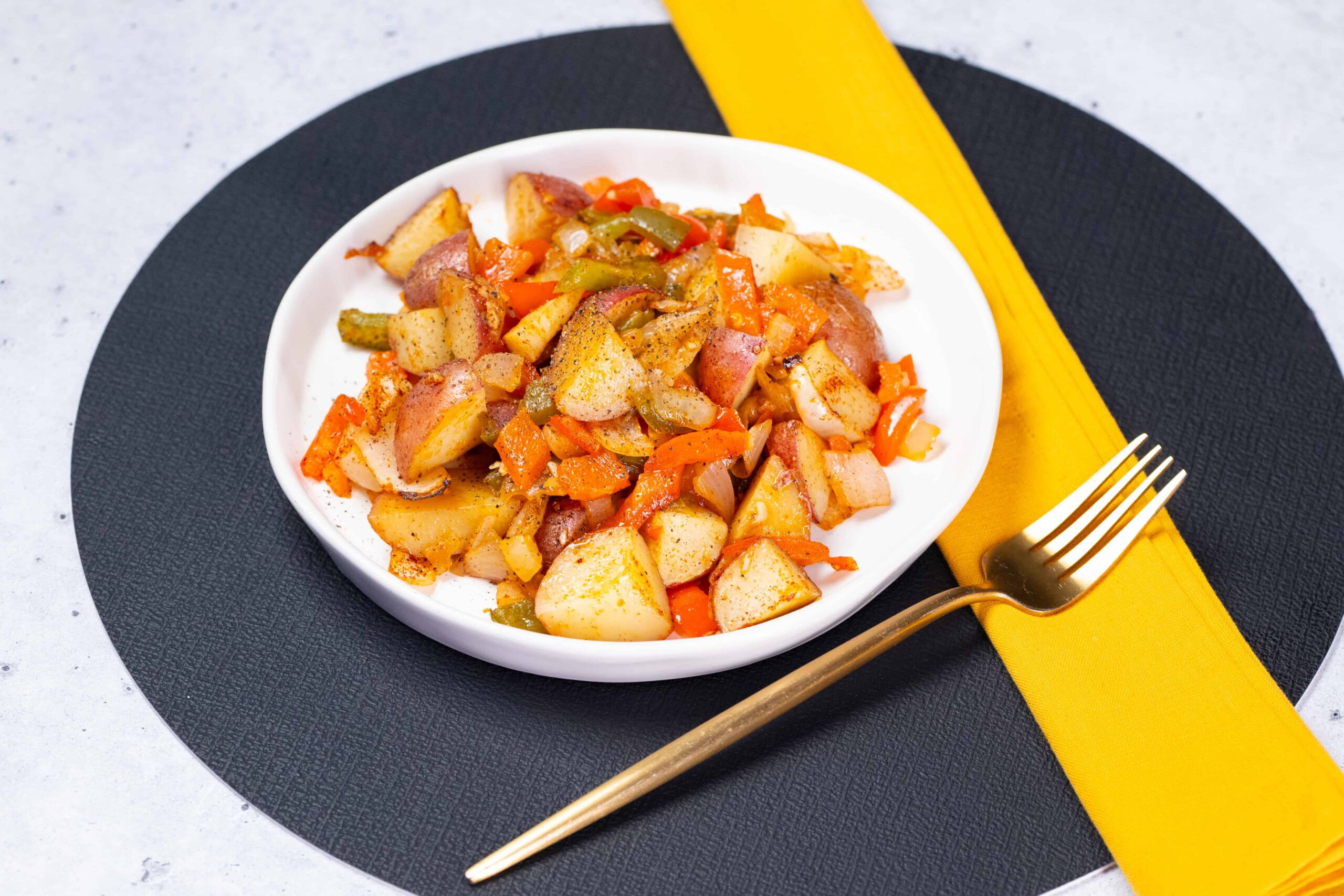 Roasted potato and vegetable hash served in a white bowl, garnished with seasoned spices, on a black round placemat with a yellow napkin and gold fork.