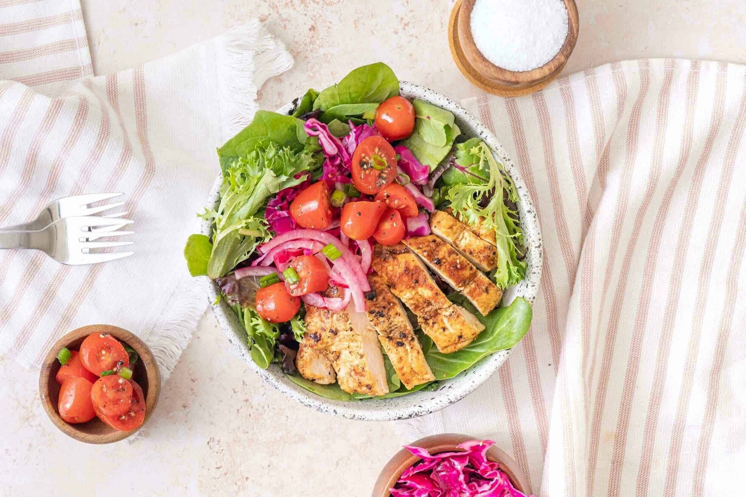 Fresh grilled chicken salad with cherry tomatoes, mixed greens, red onion, and cabbage on a rustic white plate, served with salt in a wooden bowl and a side of shredded pink cabbage, perfect for healthy eating.