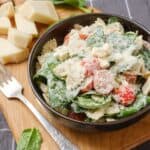Creamy pasta salad with cherry tomatoes, spinach, and grated cheese in a black bowl on wooden serving board.
