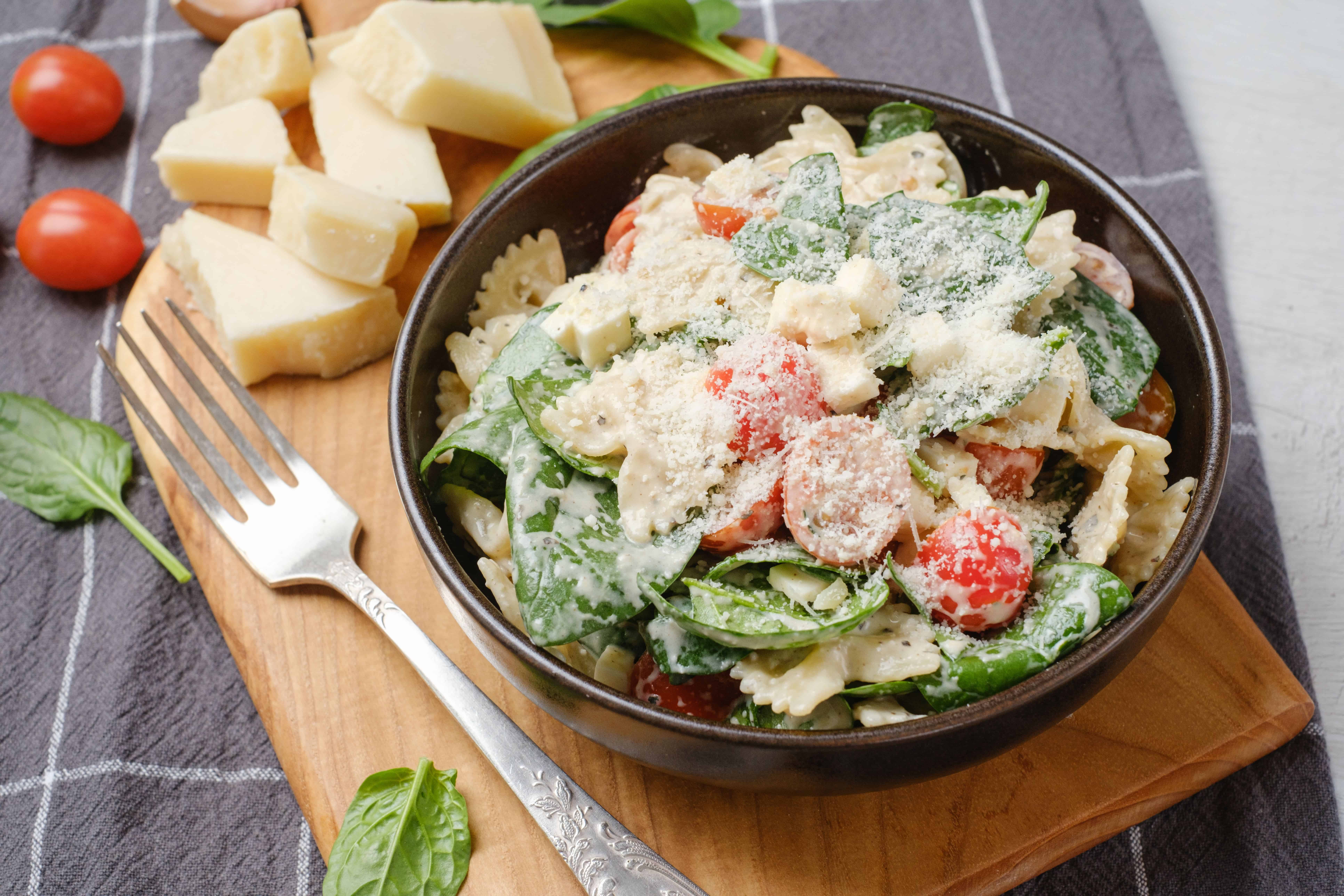 Creamy pasta salad with cherry tomatoes, spinach, and grated cheese in a black bowl on wooden serving board.