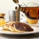 Sweet syrup being poured over a stack of fluffy pancakes with lemon slices in the background, creating a delicious breakfast scene.